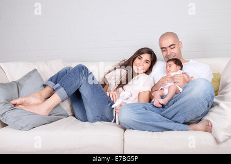 Happy family at home, cheerful young parents with cute newborn baby sitting on sofa in living room, happiness and love concept Stock Photo