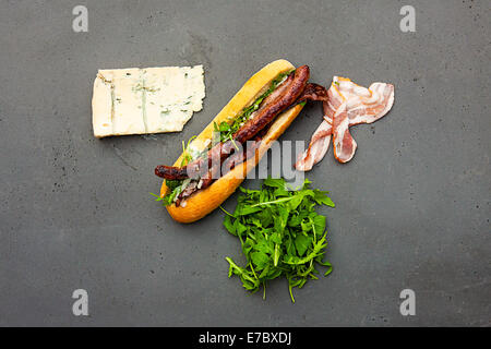 Modern hot dog with lamb sausage, bacon, rucola, gorgonzola sauce on concrete table