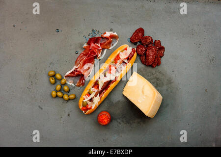 Modern hot dog with lamb sausage, prosciutto, sun-dried tomatoes, parmesan, olives, ketchup on concrete table