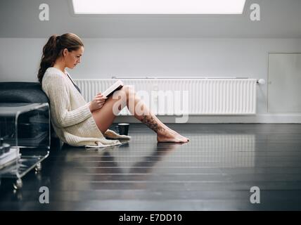 Side view of attractive young woman sitting on floor reading book. Caucasian female model at home reading a novel. Stock Photo