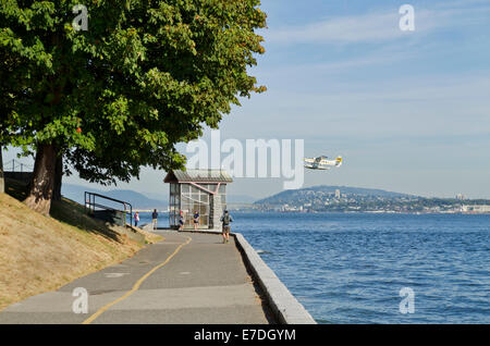 Harbour Air seaplane leaving Vancouver, flying by Stanley Park seawall.  Historic Nine o'clock Gun (encased) and tourists. Stock Photo