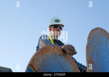 Male construction worker in hard hat and mirror sunglasses leaning against industrial machinery, low angle portrait, Fort McMurray, Alberta, Canada. Stock Photo