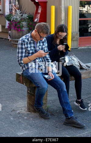 Young man and woman sitting on bench using their cell phones, Vancouver, BC, Canada Stock Photo