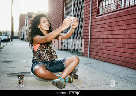 Hispanic woman taking cell phone picture on skateboard on city street Stock Photo