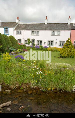 White painted English cottage with neat garden, lawn, masses of colourful spring flowers and small stream at village of Caldbeck in Cumbria Stock Photo