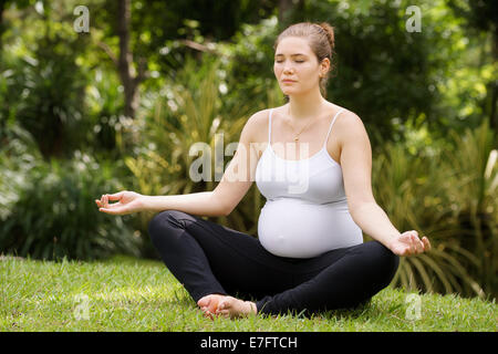 Portrait of white beautiful pregnant woman doing yoga exercise in park sitting on grass, lotus position Stock Photo