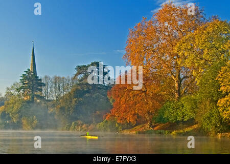 Holy Trinity church spire seen over Autumnal trees on an atmospheric misty morning, with a single scull rower on the river. Stock Photo