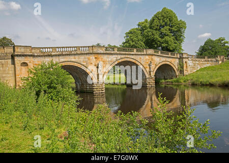 Ornate 18th century sandstone three arches bridge over River Derwent, with bridge trees and blue sky reflected in calm water Stock Photo