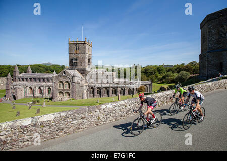 Three cyclists ride along a path by a stone church in Pembrokeshire, Wales Stock Photo