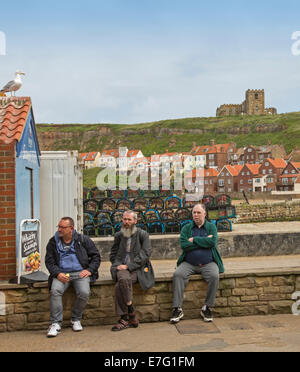 Three elderly men sitting on wall beside harbour at English seaside town of Whitby with houses on hill in background Stock Photo