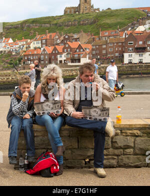 Family sitting on harbour wall eating lunch of fish and chips from takeway containers at English seaside town of Whitby Stock Photo