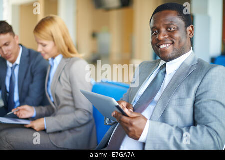 Young African-american businessman with digital tablet looking at camera in airport with two co-workers on background Stock Photo