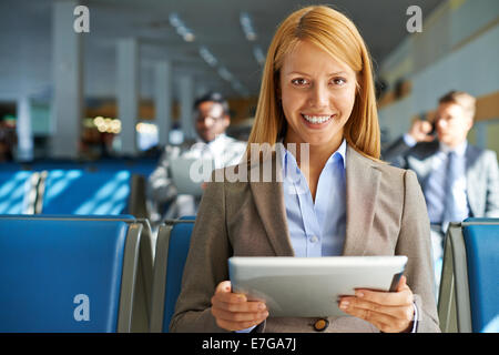 Smiling young woman with touchpad sitting in airport with two colleagues on background Stock Photo
