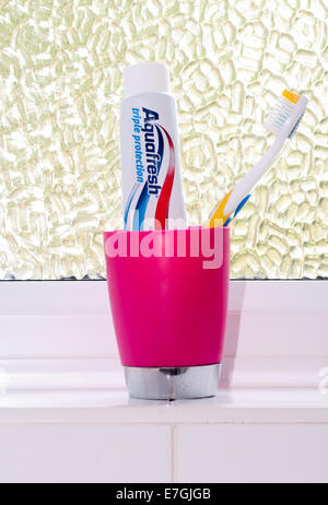 Toothbrush and Toothpaste In a Plastic Mug On a Bathroom Windowsill Stock Photo