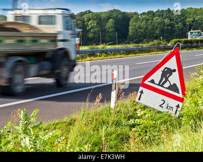 Road works ahead sign beside a busy highway. Motion blur on passing lorry. An overgrown roadworks sign, beside a road. The roadw Stock Photo