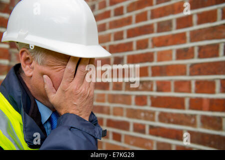 Upset building contractor, with his hands covering his face, and wearing a white safety helmet. Stock Photo
