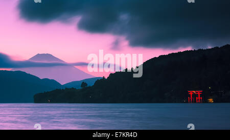 A torii gate is lit up on the shoreline, while a pink and purple sun sets over Mount Fuji in the backdrop. Stock Photo