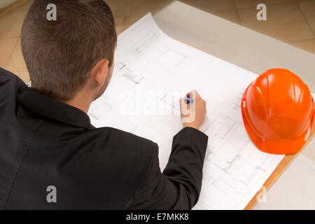 Over the shoulder view of a young architect with a pencil in his hand modifying a building plan as he sits at a table with his h Stock Photo