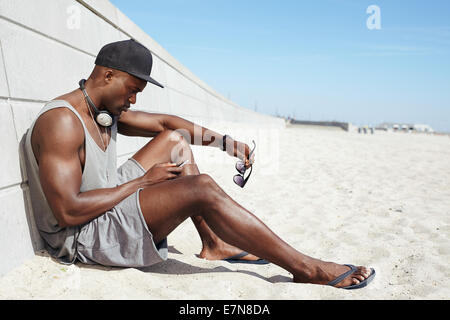 Young guy sending a text message at the beach. African man sitting on beach using mobile phone. Muscular male model outdoors. Stock Photo