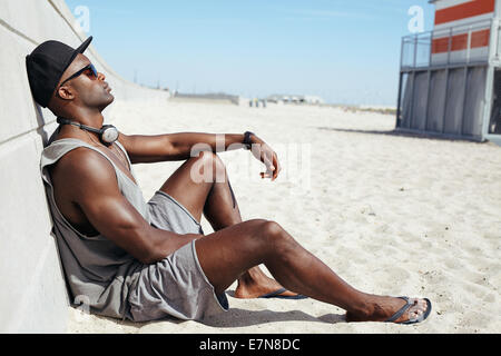Man sitting on beach leaning to a sea wall. African male model wearing sunglasses, cap and headphones relaxing on beach. Stock Photo