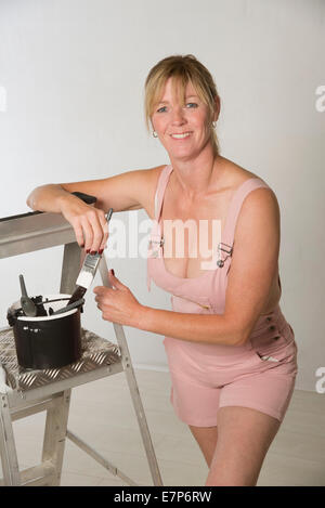 Female painter decorator wearing bib and braces overalls holding paint brush and ladder Stock Photo