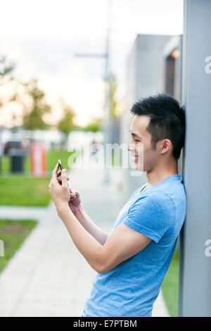 Young asian man smiling and looking at smart phone on city sidewalk Stock Photo