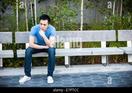 Concerned young asian man sitting on park bench with copy space Stock Photo