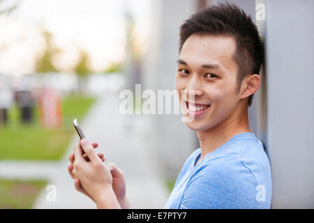 Smiling young asian man holding smart phone and looking at camera on city sidewalk Stock Photo