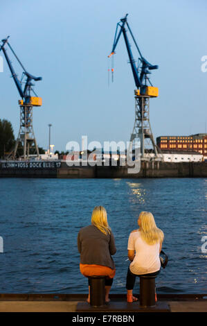 Two women sit at the port on the Elbe River with cranes in the background at dusk. Stock Photo