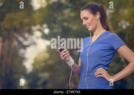 Attractive happy woman standing listening to music on a dirt track through a forest with her MP3 player and earplugs smiling