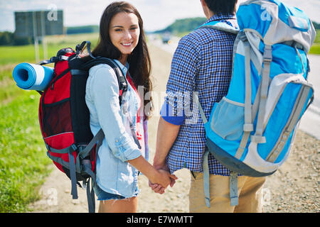 Couple of hitch-hikers standing by the road, young female looking at camera Stock Photo