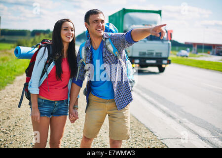 Couple of young hitch-hikers standing by highway while guy pointing somewhere Stock Photo