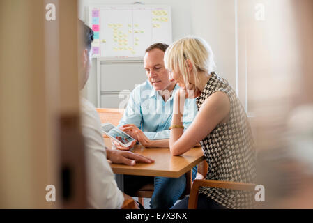 Three business colleagues in an office talking around a table and looking at a digital tablet. Stock Photo