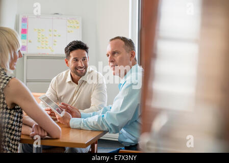 Three business colleagues in an office talking around a table and looking at a digital tablet. Stock Photo