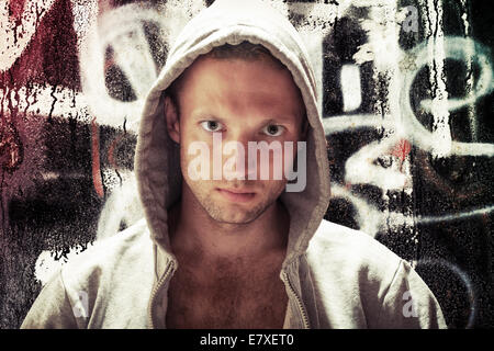 Young Caucasian man in hood, street artist portrait with grungy graffiti wall on background, toned effect