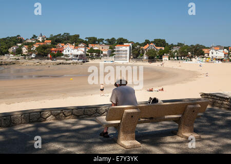 One man sitting on shady bench overlooking beach reading book. Stock Photo