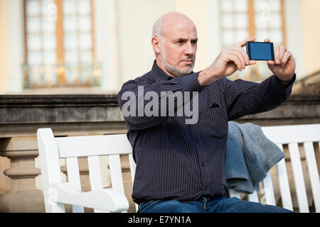 Attractive bald middle-aged man sitting on a wooden bench in an urban setting taking a photograph with his mobile phone Stock Photo