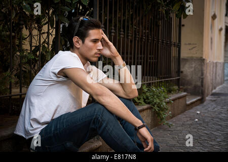 Sad, unhappy young man outdoor, sitting on pavement, holding head with his hand Stock Photo