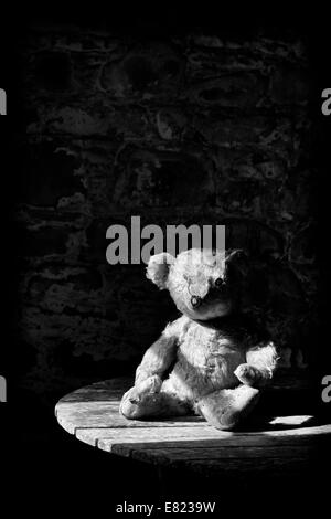 Old Threadbare One Eyed Teddy bear sat on a wooden table in sunlight. Monchrome Stock Photo