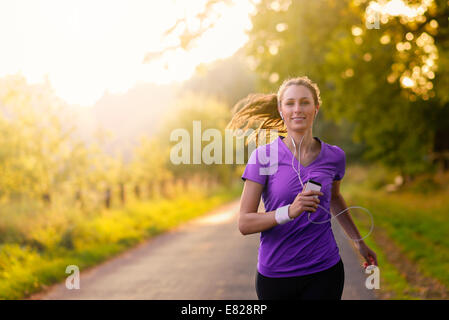 Woman listening to music on her earplugs and MP3 player while jogging along a country road in a healthy lifestyle, exercise and