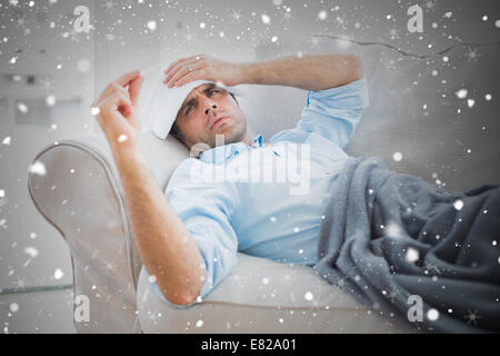 Composite image of sick man lying on sofa checking his temperature under a blanket Stock Photo