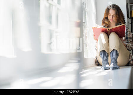 A woman seated by a window, reading with a book on her lap. Stock Photo