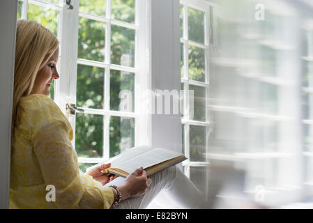 Woman sitting by a window, reading a book. Stock Photo