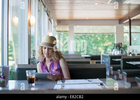 A woman in a hat sitting in a diner, holding a digital tablet, looking at the screen. Stock Photo