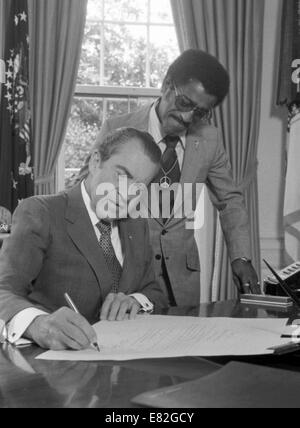 President Richard Nixon signing a document at his desk in the Oval Office as Sammy Davis, Jr., looks on. July 1, 1971 Stock Photo