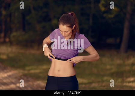 Woman athlete checking her pulse belt during a training run through woodland lifting her top to expose her toned slender midriff Stock Photo