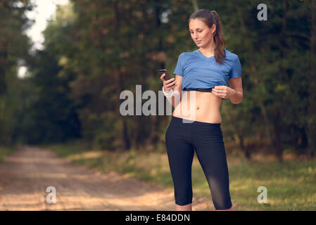 Young athletic woman out training in the woods standing looking at her smart phone as she uses a mobile application electronical Stock Photo