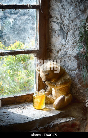 Threadbare One Eyed Teddy bear on an old window ledge looking at a pot of honey Stock Photo