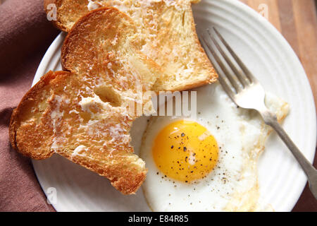 Two slices of buttered toast with a fried egg on a plate with a fork Stock Photo