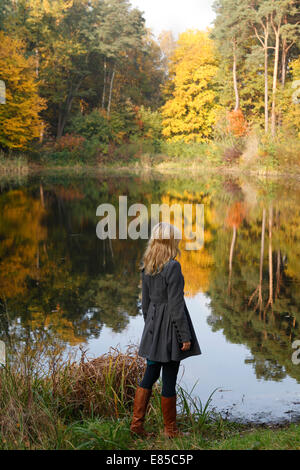 a young dreamy blonde woman is standing on the bank of an autumn lake reflecting the stunning fall colours of the trees (model-release available) Stock Photo